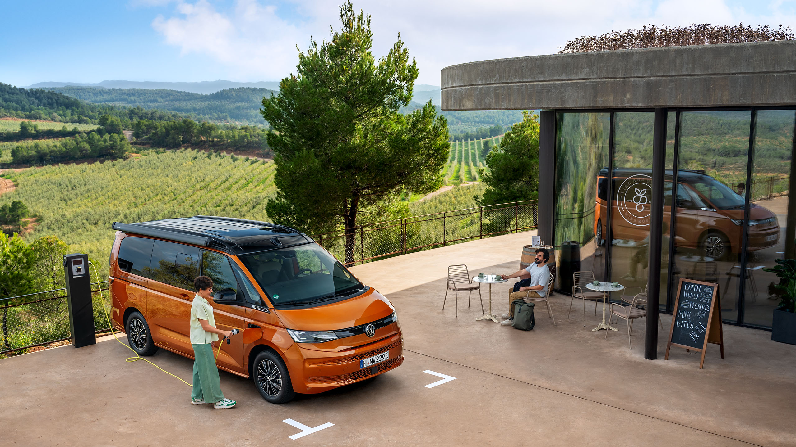 Ein VW California parkt neben einem Cafe und wird an einer Ladestadion geladen.